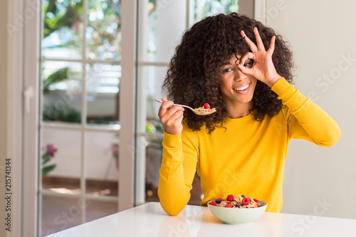 African american woman eating cereals, raspberries and blueberries with happy face smiling doing ok sign with hand on eye looking through fingers
