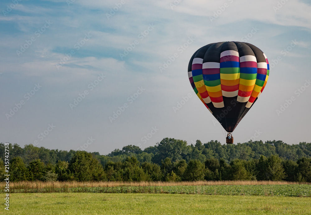 Naklejka premium Red, Yellow, Orange, Green, and White Teardrop Shaped Hot Air Balloon Over Corn Field on Sunny Day with Trees and Cloudy Blue Sky in Background