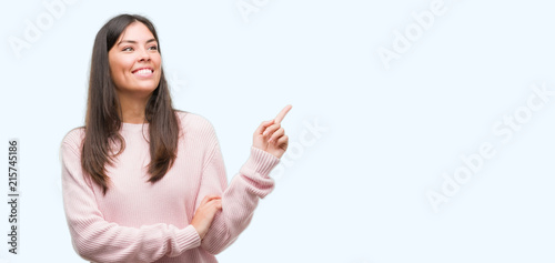 Young beautiful hispanic woman wearing a sweater with a big smile on face, pointing with hand and finger to the side looking at the camera.
