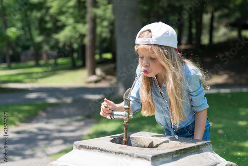 A little cute blonde girl is drinking from Water Fountain in a park. She is wearing a jean t-shirt and a baseball cap