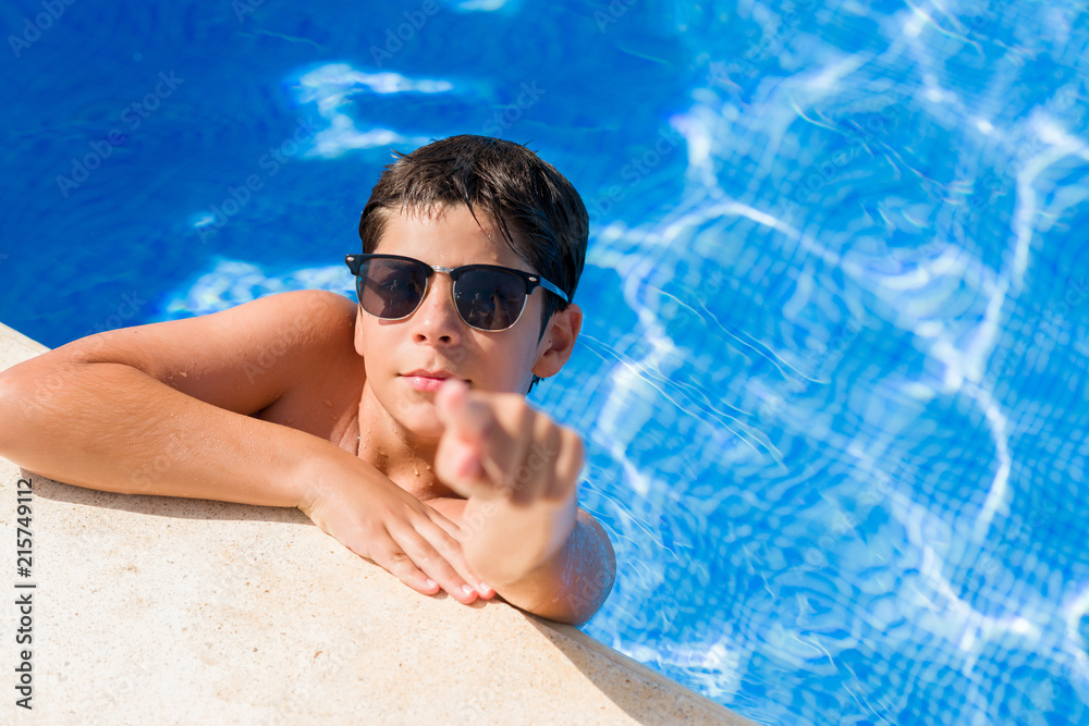 Young child on holiday at the swimming pool by the beach pointing with finger to the camera and to you, hand sign, positive and confident gesture from the front