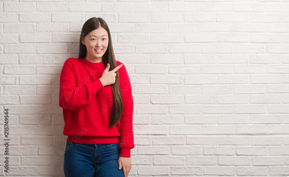 Young Chinese woman over brick wall cheerful with a smile of face pointing with hand and finger up to the side with happy and natural expression on face looking at the camera. Young Chinese woman over brick wall cheerful with a smile of face pointing with hand and finger up to the side with happy and natural expression on face looking at the camera.