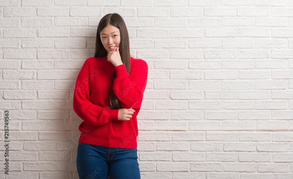Young Chinese woman over brick wall looking confident at the camera with smile with crossed arms and hand raised on chin. Thinking positive. Young Chinese woman over brick wall looking confident at the camera with smile with crossed arms and hand raised on chin. Thinking positive.