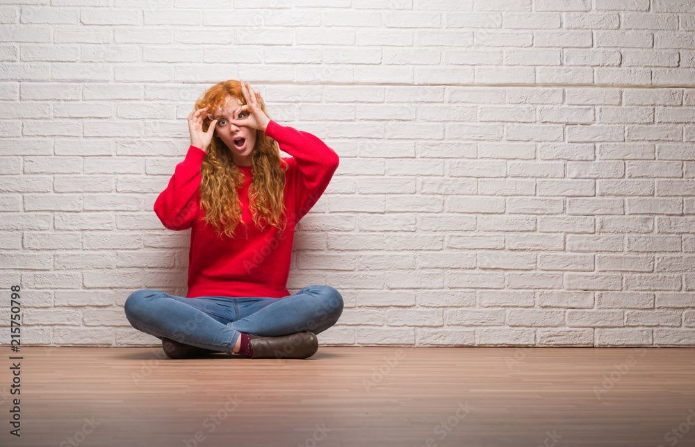 Young redhead woman sitting over brick wall doing ok gesture shocked with surprised face, eye looking through fingers. Unbelieving expression. Young redhead woman sitting over brick wall doing ok gesture shocked with surprised face, eye looking through fingers. Unbelieving expression.