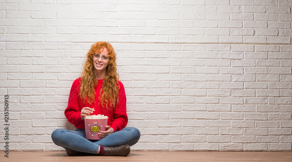 Young redhead woman sitting over brick wall eating popcorn with a happy face standing and smiling with a confident smile showing teeth Young redhead woman sitting over brick wall eating popcorn with a happy face standing and smiling with a confident smile showing teeth
