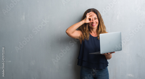 Middle age hispanic woman standing over grey grunge wall using laptop with happy face smiling doing ok sign with hand on eye looking through fingers