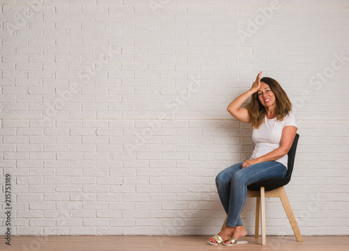 Middle age hispanic woman sitting on chair over white brick walll stressed with hand on head, shocked with shame and surprise face, angry and frustrated. Fear and upset for mistake.