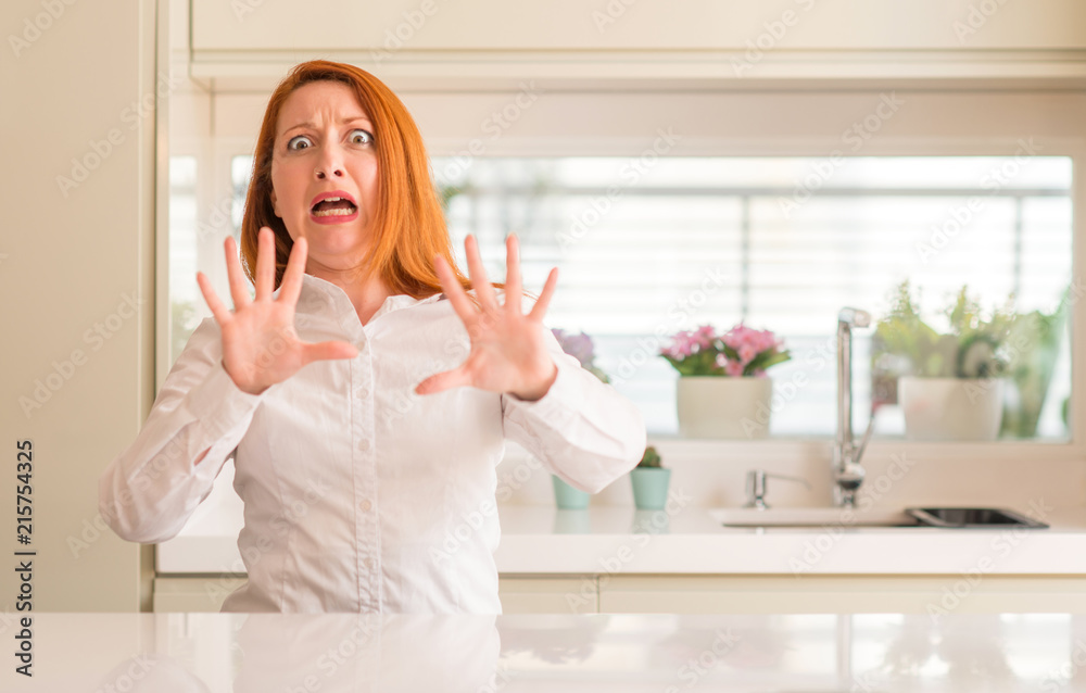 Redhead woman at kitchen afraid and terrified with fear expression stop gesture with hands, shouting in shock. Panic concept. Redhead woman at kitchen afraid and terrified with fear expression stop gesture with hands, shouting in shock. Panic concept.