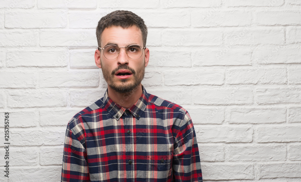Young adult man standing over white brick wall scared in shock with a surprise face, afraid and excited with fear expression Young adult man standing over white brick wall scared in shock with a surprise face, afraid and excited with fear expression