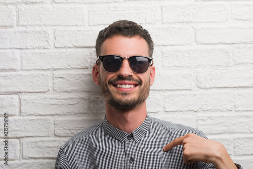 Young adult man wearing sunglasses standing over white brick wall with surprise face pointing finger to himself photo