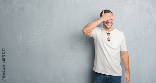 Young caucasian man over grey grunge wall wearing sunglasses smiling and laughing with hand on face covering eyes for surprise. Blind concept.