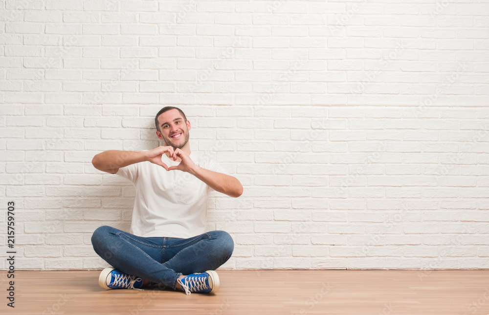 Young caucasian man sitting on the floor over white brick wall smiling in love showing heart symbol and shape with hands. Romantic concept.