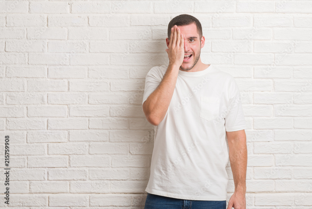 Young caucasian man standing over white brick wall covering one eye with hand with confident smile on face and surprise emotion. Young caucasian man standing over white brick wall covering one eye with hand with confident smile on face and surprise emotion.
