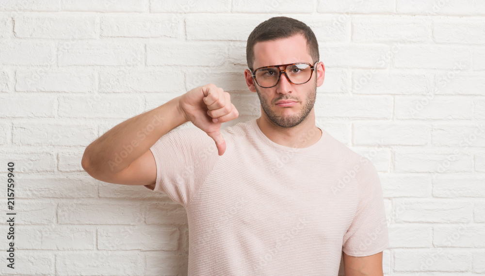 Young caucasian man standing over white brick wall wearing glasses with angry face, negative sign showing dislike with thumbs down, rejection concept Young caucasian man standing over white brick wall wearing glasses with angry face, negative sign showing dislike with thumbs down, rejection concept