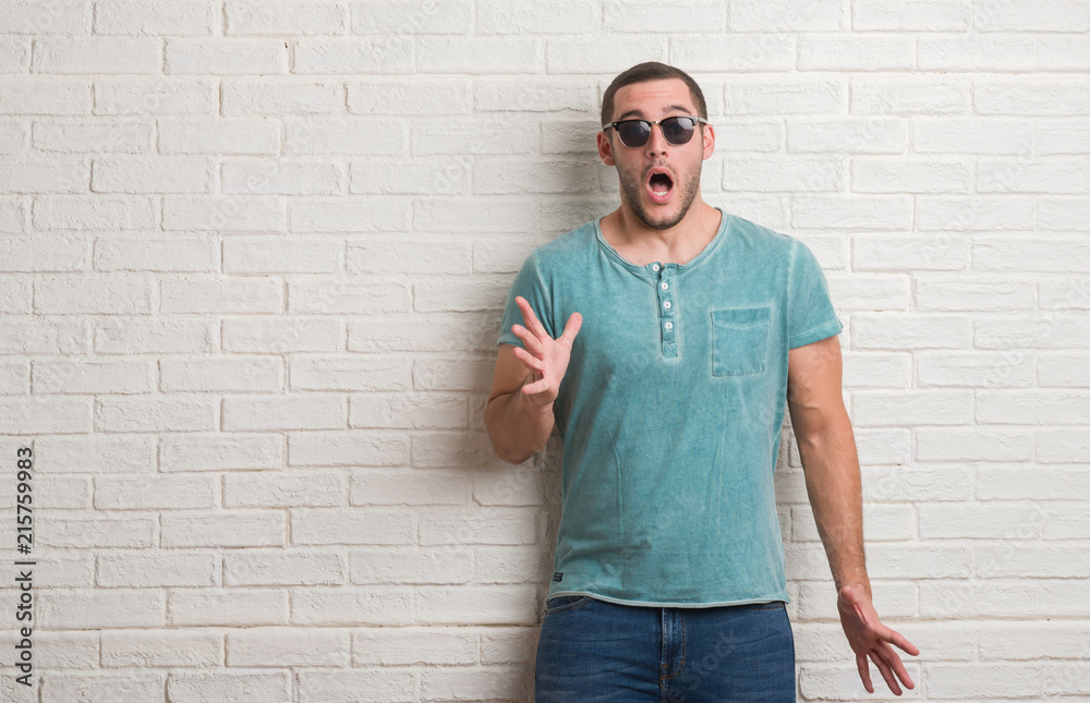 Young caucasian man standing over white brick wall wearing sunglasses scared in shock with a surprise face, afraid and excited with fear expression Young caucasian man standing over white brick wall wearing sunglasses scared in shock with a surprise face, afraid and excited with fear expression