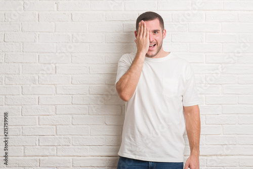 Young caucasian man standing over white brick wall covering one eye with hand with confident smile on face and surprise emotion.