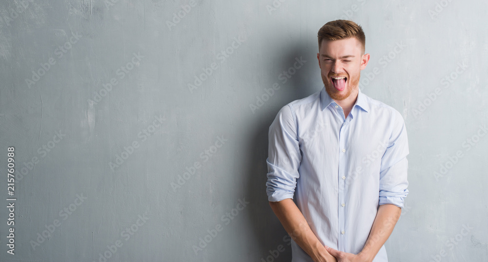 Young redhead business man over grey grunge wall sticking tongue out happy with funny expression. Emotion concept.