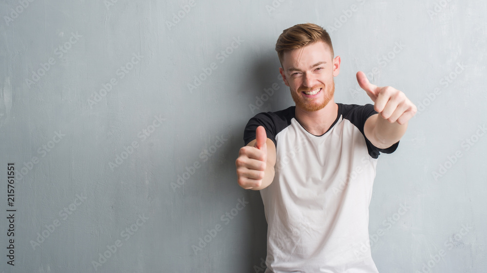 Young redhead man over grey grunge wall approving doing positive gesture with hand, thumbs up smiling and happy for success. Looking at the camera, winner gesture. - obrazy, fototapety, plakaty 