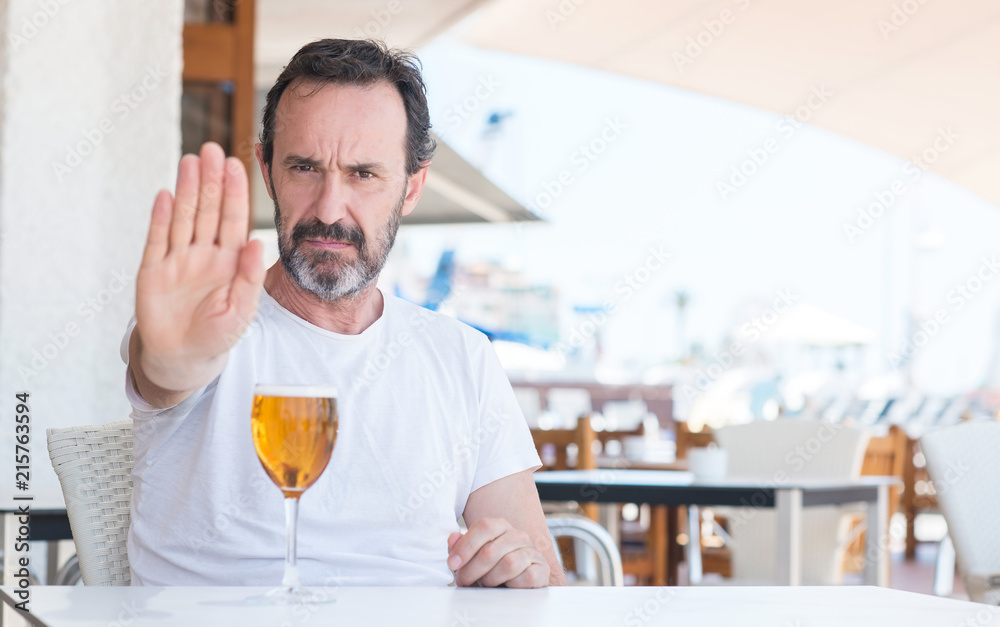Handsome senior man drinking beer at restaurant with open hand doing stop sign with serious and confident expression, defense gesture Handsome senior man drinking beer at restaurant with open hand doing stop sign with serious and confident expression, defense gesture