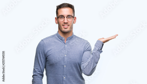 Handsome young elegant man wearing glasses smiling cheerful presenting and pointing with palm of hand looking at the camera.