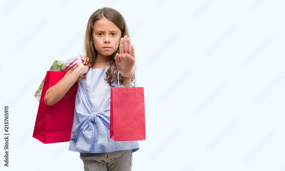 Brunette hispanic girl holding shopping bags with open hand doing stop sign with serious and confident expression, defense gesture Brunette hispanic girl holding shopping bags with open hand doing stop sign with serious and confident expression, defense gesture
