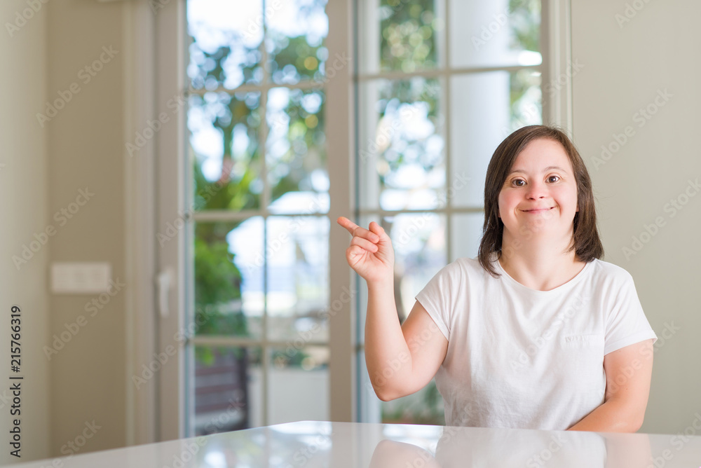 Down syndrome woman at home with a big smile on face, pointing with hand and finger to the side looking at the camera.