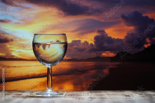 A glass of drinking water is on a wooden table against the background of the sea sunset.