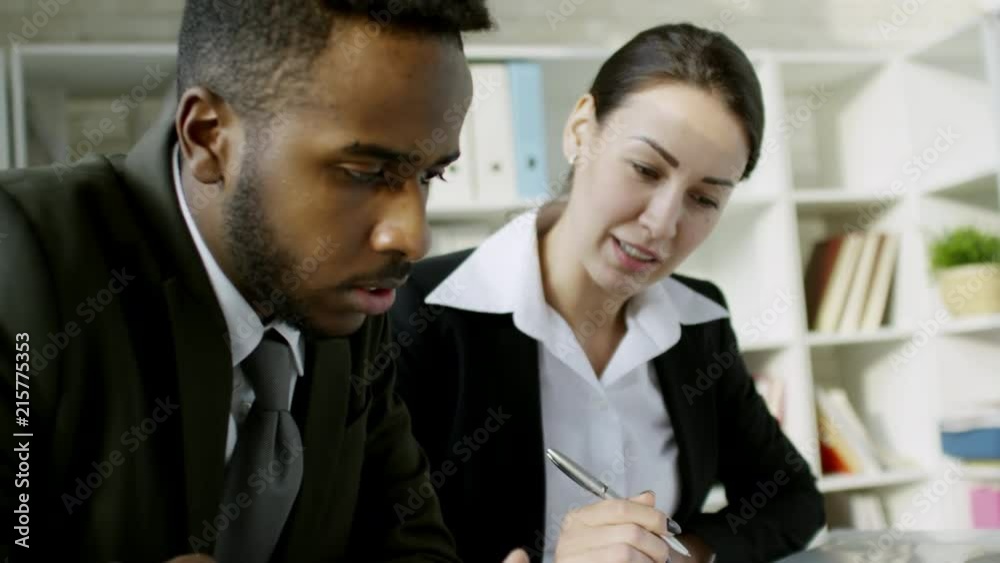 Handheld shot of two multiethnic business partners sitting at desk in office and discussing documents Handheld shot of two multiethnic business partners sitting at desk in office and discussing documents