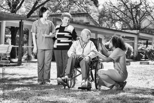 Two female doctors helping elderly man and woman with physical disability ih rehab facility garden