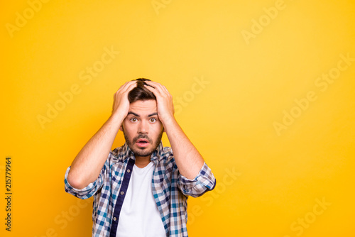 Portrait of frightened brunette man in a plaid shirt holds his hands behind his head and looks into the camera isolated on shine yellow background with copy space for text