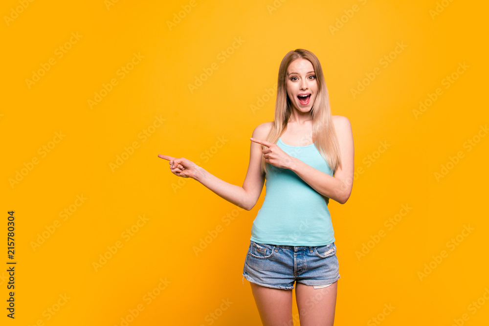 Attractive cute straight-haired blonde caucasian smiling girl, wearing  casual blue shirt and jeans shorts, amazed, pointing showing with two  fingers to object. Isolated over yellow background Stock Photo | Adobe Stock