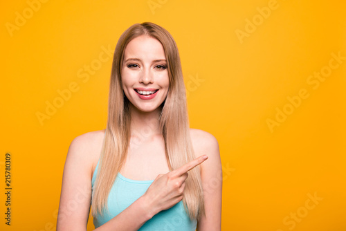 Advertisement concept. Confident young blond woman with beaming smile pointing with her index finger on copy space and looking on camera isolated on shine yellow background