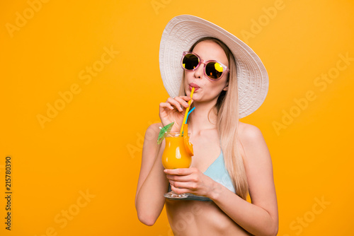 Attractive nice cute straight-haired beautiful blonde caucasian girl, wearing blue swimsuit, sun glasses, hat. Glad, drinking glass of orange cocktail. Isolated over yellow background