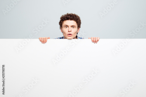 Brunet curly hair man standing behind the white blank banner and hald blank white copy space isolated on light gray background gaze into camera photo