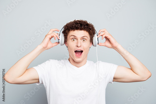 WOW! Young guy listening good music in big white headphones with wide open mouth and eyes isolated on light gray background
