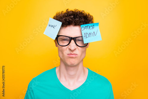 Handsome curly-haired upset young guy wearing casual green t-shirt and glasses. Two stickers with texts pinned on head. Isolated over yellow background photo