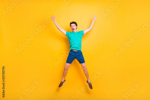 Full size length body picture of handsome curly-haired funny young guy wearing casual green t-shirt, shorts, shoes, jumping in air, hands up, star figure. Isolated over yellow background