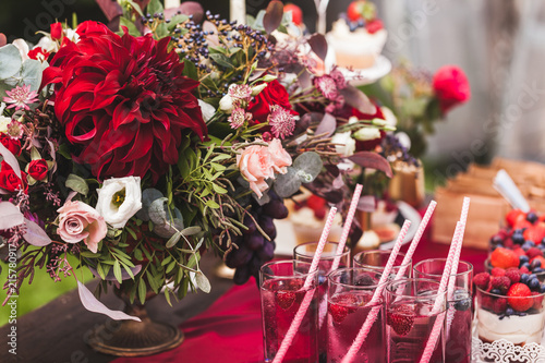 Wedding reception table decorated with red flowers, sweets, drinks and silk. Rustic style