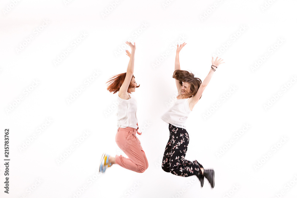 Two happy and positive women jump over a white wall in studio - obrazy, fototapety, plakaty 