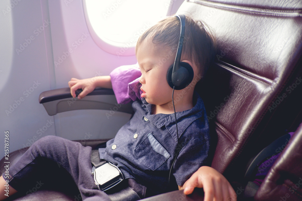 Naklejka premium Cute little Asian 24 months / 2 years old toddler baby boy child sleeping on Airplane, Toddler boy sitting with safety belt on wearing headphones while traveling in airplane, Kids Fly Safe concept