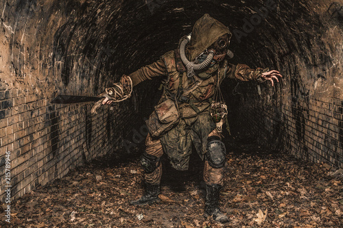Post apocalyptic mutant creature or survivor in tatters and gas mask jumps out of darkness and attacking with handmade machete in abandoned tunnel, frightening dungeon or city old sewage collector photo