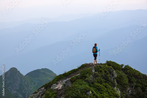 The girl with the touristic equipment goes up to the peak of the rocky high hill with the lawn. The scenery of the mountains in the fog.