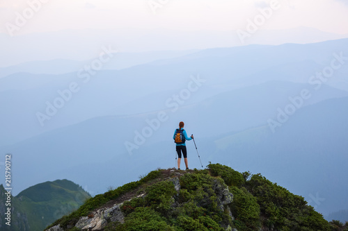 The girl with the touristic equipment goes up to the peak of the rocky high hill with the lawn. The scenery of the mountains in the fog. © Vitalii_Mamchuk