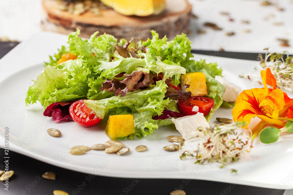 European Italian salad of lettuce, cherry tomatoes, pumpkin, edible pumpkin flowers, and pumpkin seeds, in a white plate. Copy space, selective focus European Italian salad of lettuce, cherry tomatoes, pumpkin, edible pumpkin flowers, and pumpkin seeds, in a white plate. Copy space, selective focus