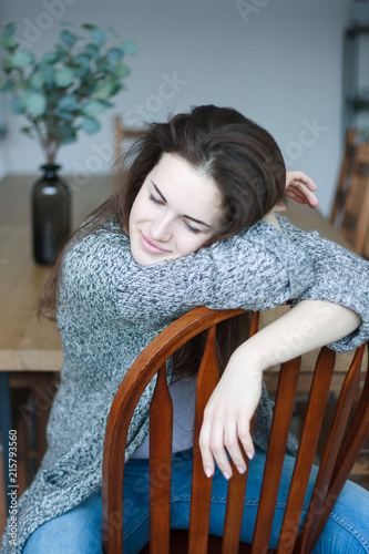 Beautiful young brunette woman sitting on the wooden chair, wearind jeans and a sweater, looking down and smiling. photo