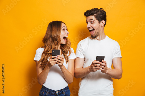 Photo of positive excited people man and woman screaming and looking at each other while both using mobile phones, isolated over yellow background photo