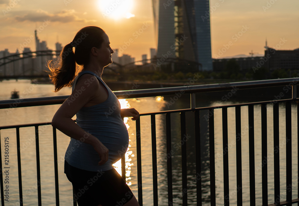 Fototapeta premium Active pregnant woman running across the bridge with a beautiful view of the city of Frankfurt in Germany