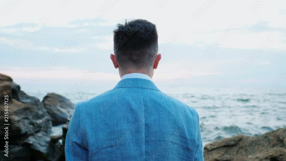 Portrait of young attractive business man or groom stands on sea shore with rocks and watching beautiful sunset or sunrise alone. 4k. Slow motion Portrait of young attractive business man or groom stands on sea shore with rocks and watching beautiful sunset or sunrise alone. 4k. Slow motion