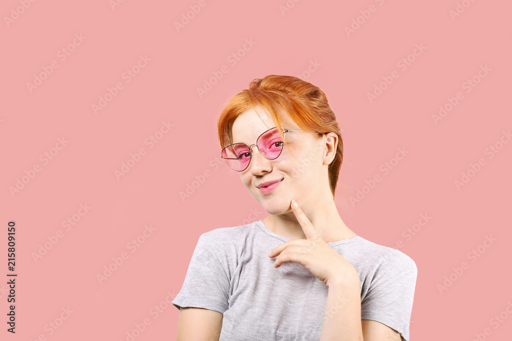 Attractive young woman with long red hair, wearing pink cat eye sunglasses, showing flirtatious body language, touching face. Beautiful redhead female flirty facial expression. Background, copy space. Attractive young woman with long red hair, wearing pink cat eye sunglasses, showing flirtatious body language, touching face. Beautiful redhead female flirty facial expression. Background, copy space.