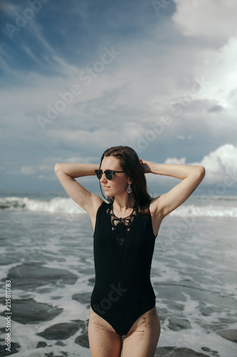 Happy smile brunette Woman wearing black swimsuit at beautiful sky and ocean background on beach with black sand